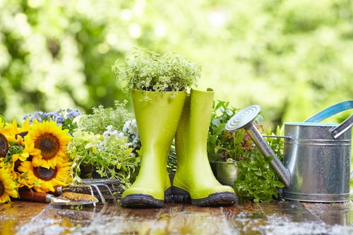 The image depicts a gardening scene set outdoors with a blurred green background. In the foreground, a rustic wooden table holds a variety of gardening tools and decorative elements. There are bright yellow sunflower heads, white and purple flowering plants, and foliage arranged in a loose, natural style. Prominently, a pair of lime green rubber rain boots, filled with white flowering plants, are positioned upright on the table, adding a whimsical touch. To the right of the boots, there is a metal watering can with a long spout, reflecting sunlight, and additional green potted plants nearby. The scene suggests a well-maintained garden environment suitable for gardening or landscaping services, with features typical of backyard or patio gardens that might be serviced by companies like Gardening Services Walthamstow near the E17 postcode area. The lighting is natural, indicating a sunny day, and the overall composition emphasizes outdoor plant care and garden decoration elements typical of professional gardening work in the Walthamstow locality.