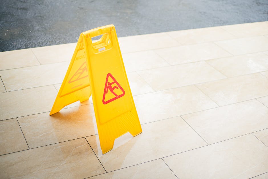 A bright yellow plastic warning sign with a red triangular symbol indicating a wet or slippery floor is positioned on beige tiled flooring near the entrance of an outdoor garden. The tiles have a smoo